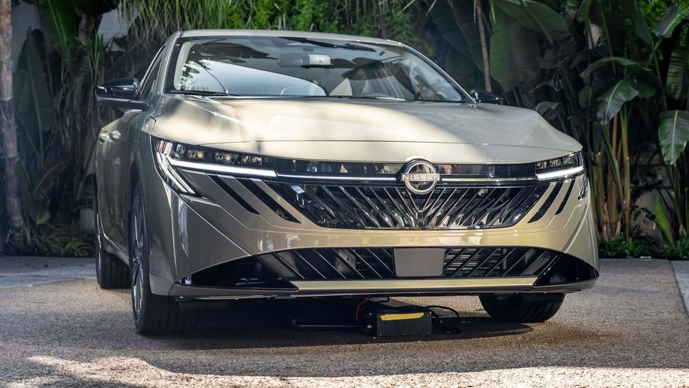 A low-angle, front-view shot of a modern, light-gold or champagne-colored Nissan Sentra sedan, showcasing its bold, angular V-motion grille with intricate detailing, slim horizontal LED headlights, and the prominent Nissan logo. The car is parked outdoors with lush greenery in the background.