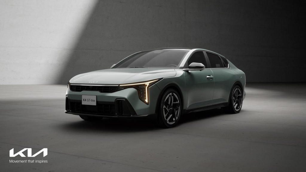 
A low-angle studio shot of the Kia K4 GT-Line in a light greenish-gray color, featuring its angular headlights and modern sedan profile. The Kia logo and "Movement that inspires" slogan are visible in the lower left corner.
