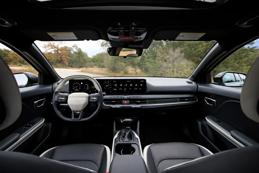 
A centered view of the interior dashboard of the Kia K4, showing the wide digital display panel, a two-tone black and light gray steering wheel, and the center console with the gear selector. The black leather seats with white accents are visible in the foreground, and a road with trees can be seen through the windshield.