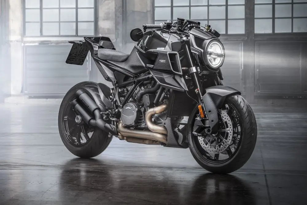 A matte black Brabus 1300 R motorcycle, a high-performance naked bike, showcasing its carbon fiber body panels, custom exhaust, and single round headlight in an indoor industrial setting.