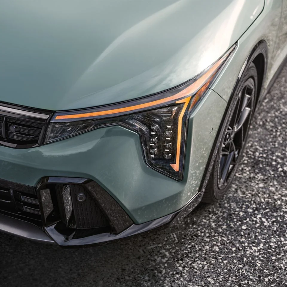 
A close-up, elevated view of the front driver's side of the Kia K4 in a light green color, focusing on the aggressive, vertically-oriented LED headlight cluster with the amber daytime running light illuminated, and the black lower air intake. The car is parked on a gravel surface.