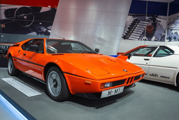 
An iconic bright orange BMW M1, a classic example of Forgotten Supercars, is on display indoors, likely at a museum. The mid-engine sports car features its distinctive angular design and black trim, parked next to a white M1 model. The background shows a graphic display related to its history.