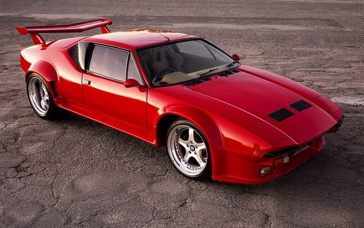 A stunning, high-angle shot of a bright red De Tomaso Pantera, a classic example of Forgotten Supercars, is parked on dark asphalt. This highly modified version features aggressive wide-body fender flares, a large rear wing, and polished multi-spoke wheels.
