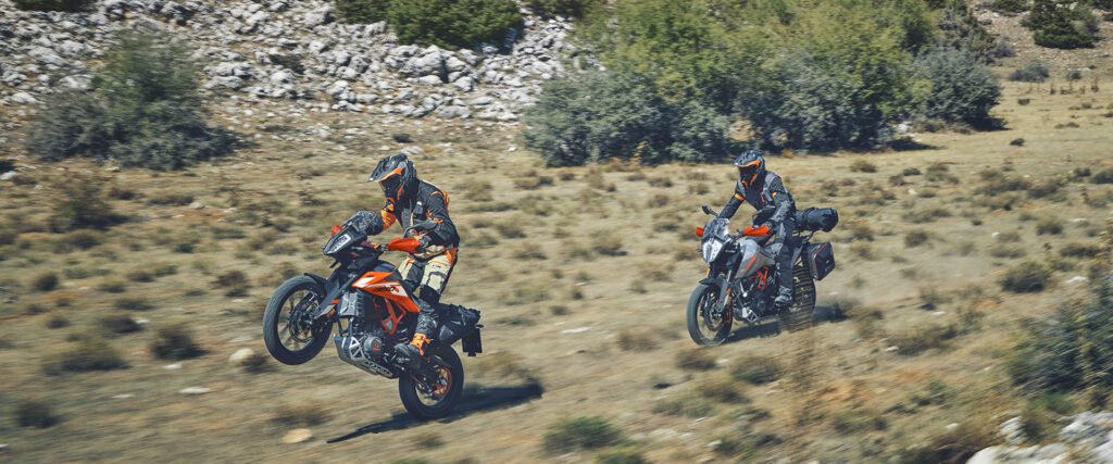 Two riders on KTM Adventure 390 motorcycles are off-roading on a dry, rocky, and grassy trail, with the rider in the foreground performing a wheelie. The focus keyword is "KTM Adventure 390".