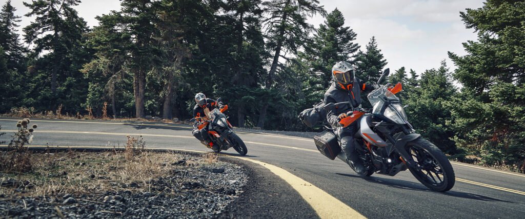 Two motorcyclists on KTM Adventure 390 bikes lean into a curve on a paved, winding mountain road, surrounded by tall pine trees. The focus keyword is "KTM Adventure 390".