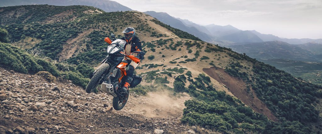 A single rider on a KTM Adventure 390 motorcycle is captured mid-air, jumping or wheelieing up a steep, rocky, dirt hill in a mountainous landscape. The focus keyword is "KTM Adventure 390".
