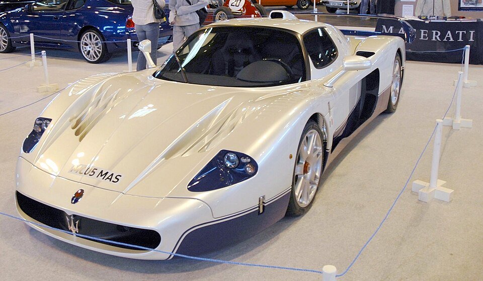 
A high-angle shot of a stunning white and blue Forgotten Supercars model, the Maserati MC12, displayed indoors at an auto show. The ultra-rare coupe features an aggressive, long body, a prominent front badge, and a racing livery stripe. Other cars and people are visible in the background.