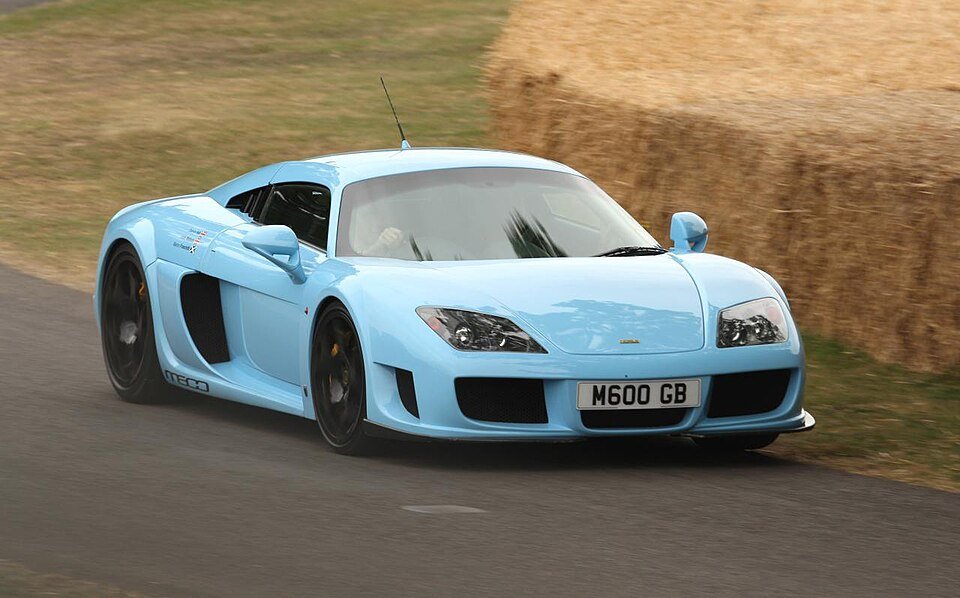 
A light blue Noble M600, a British example of Forgotten Supercars, is captured in motion on an asphalt road. The mid-engine sports car features large side air intakes and black wheels, with a blurred background showing straw bales and a dry field.