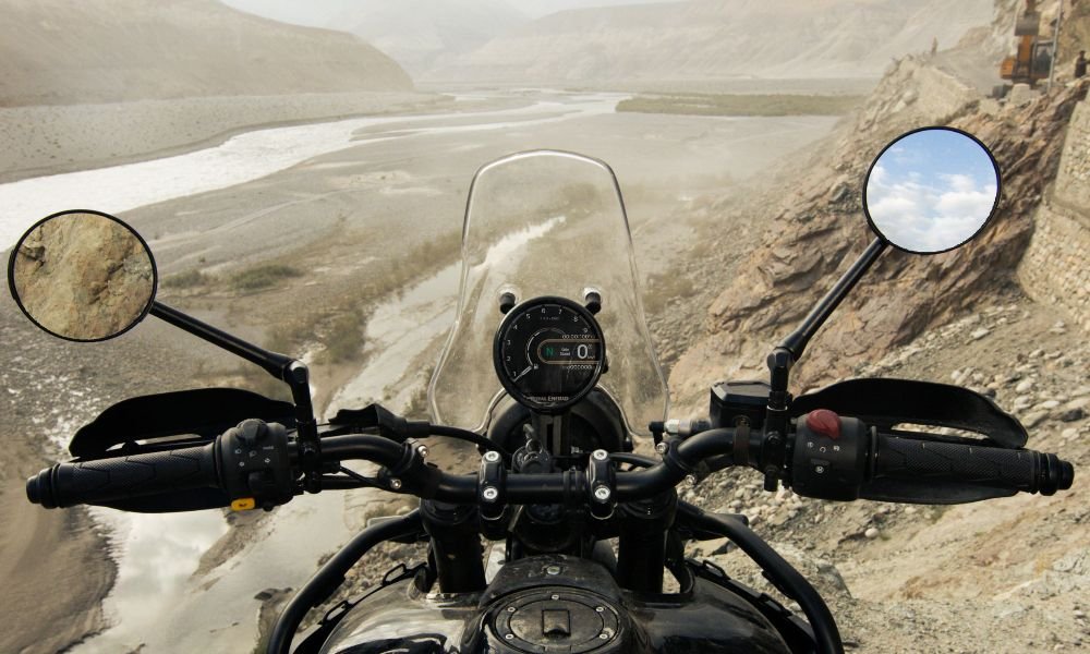 Cockpit view from a Himalayan 452 motorcycle, showing the handlebars, windshield, and a round digital instrument cluster. The view ahead is a vast, dry river valley with surrounding mountains. The focus keyword is "Himalayan 452".