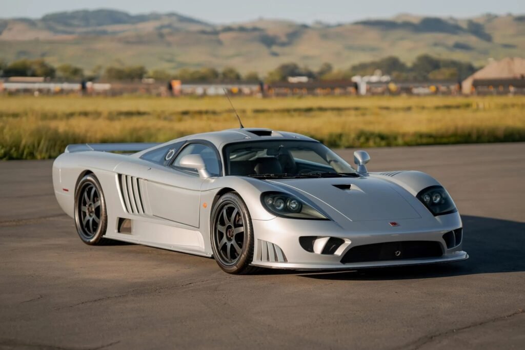 
A striking silver Forgotten Supercars model, the Saleen S7, is pictured on an asphalt surface with a golden field and rolling hills in the background under a clear sky. The low-slung American supercar features aggressive styling, large side vents, and multi-spoke black wheels.