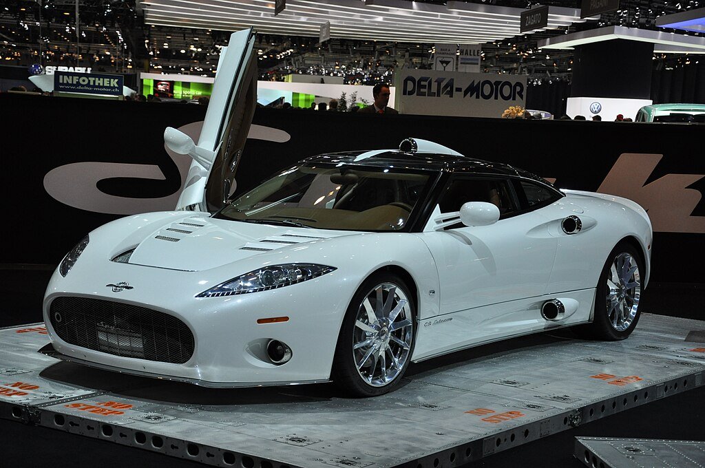 
A glossy white Spyker C8 Aileron, an example of a Forgotten Supercars, is prominently displayed on a platform at an auto show. The Dutch sports car features a distinctive front grille, chrome turbine-style wheels, exposed side exhausts, and its signature scissor door open on the driver's side. The background shows the bustling show floor.