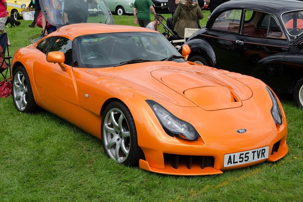 
A close-up of a vibrant orange TVR Sagaris, an aggressively styled British Forgotten Supercars model, is parked on a grassy field at a show. The car features distinctive front air intakes, clear headlight covers, and silver alloy wheels. Other classic cars and people are visible in the background.
