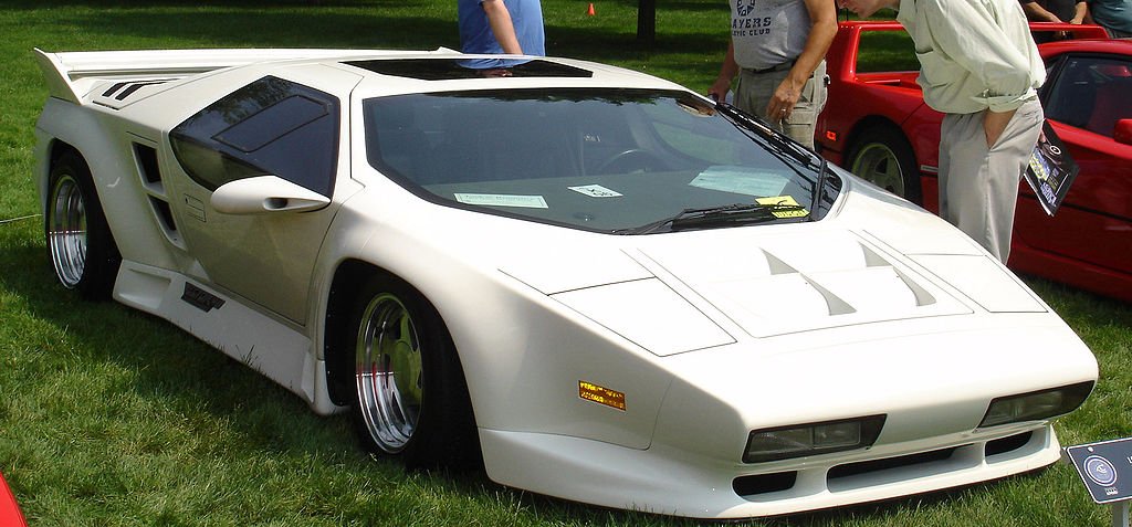 
A striking white Forgotten Supercars model, the Vector W8, is shown parked on a grassy display area. The car has an aggressive, wedge-shaped design with a large rear wing and blacked-out windows. People are visible standing near the car in the background.