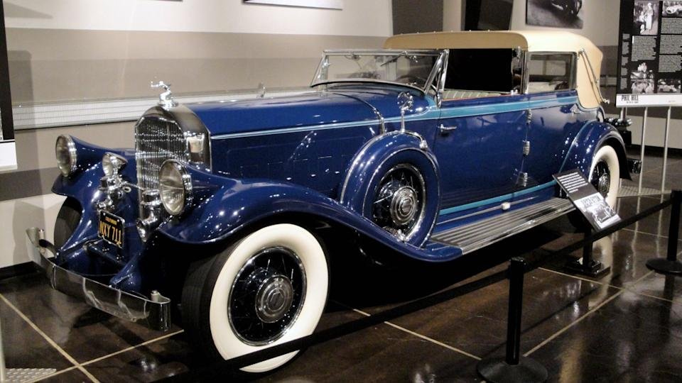 
An ornate, dark blue classic convertible with wide whitewall tires on display in a museum, representing the luxury gasoline-powered vehicles that dominated the market following the initial era of electric car history in the early 20th century.