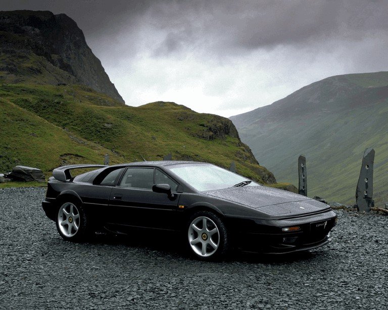 
A moody, side profile shot of a black Lotus Esprit V8, one of the classic Forgotten Supercars, is parked on a dark gravel surface. The car has a distinctive wedge shape, a rear wing, and silver alloy wheels. The background features dramatic, green-sloped mountains under a cloudy, overcast sky.